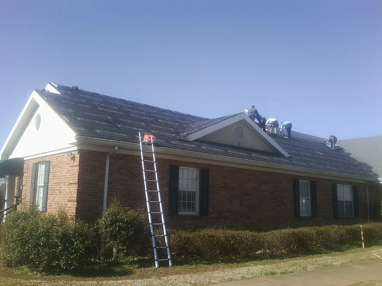 Crew tearing shingles off a brick ranch roof with a ladder set — Metal Roof Repair work by our crew in Mc Knightstown