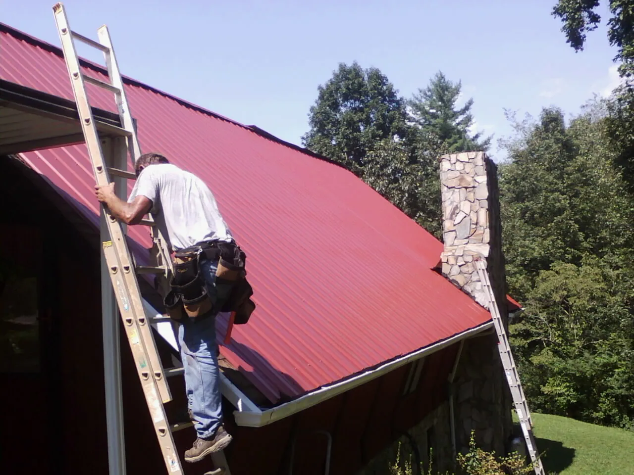 Roofer on a ladder beside a red metal roof and stone chimney — Soffit & Fascia Repair by Roofing Brothers in Mc Knightstown